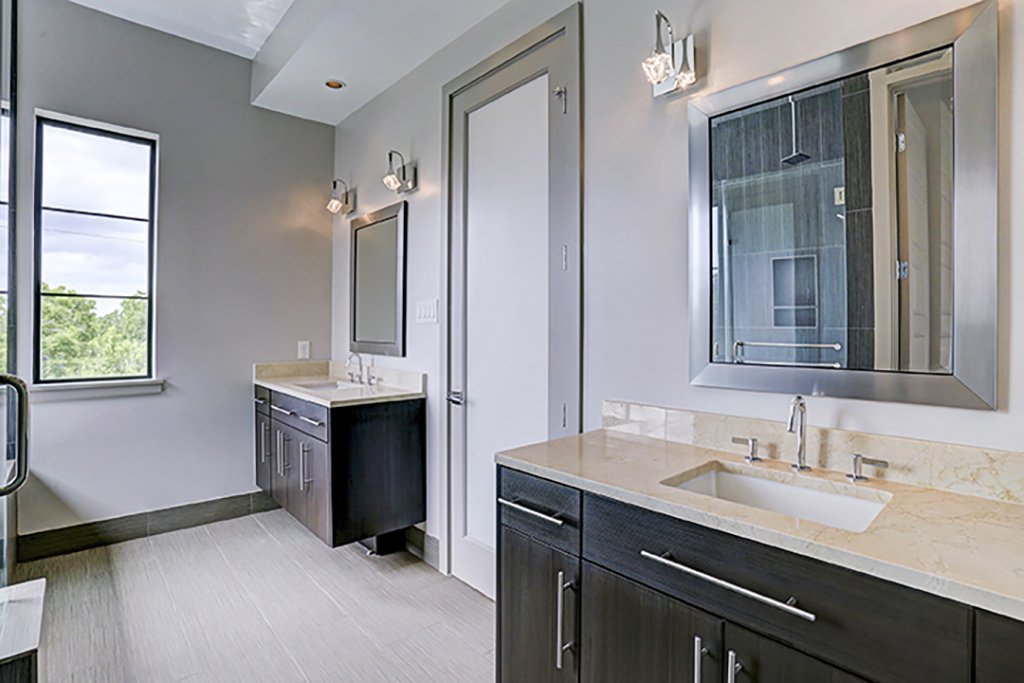 Bathroom with two sinks, dark cabinets, marble countertops, wall-mounted mirrors, and a window showing greenery outside.