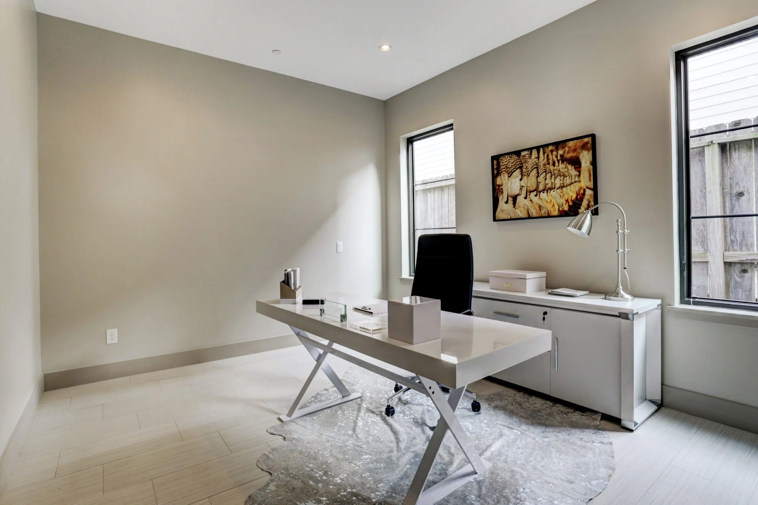 Minimalist office with white desk, black office chair, white cabinet, framed art of Buddha statues, two windows, and a silver desk lamp.