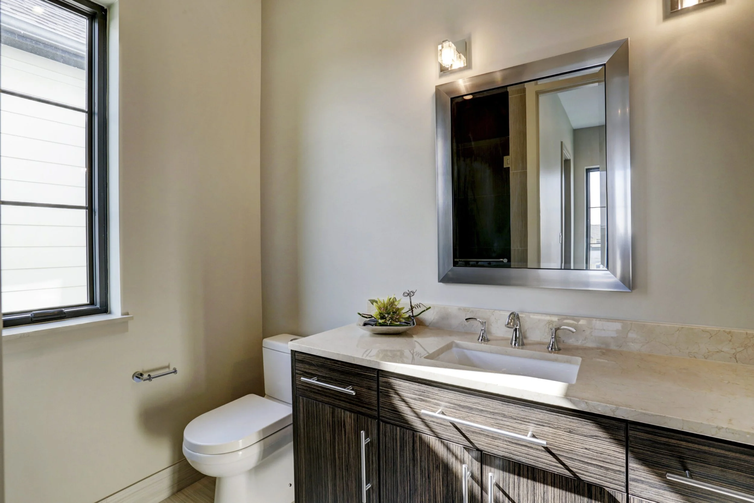 A modern bathroom with a large mirror, a marble countertop with a sink, a dark wood vanity, a small plant on the counter, a window with black trim, and a white toilet.