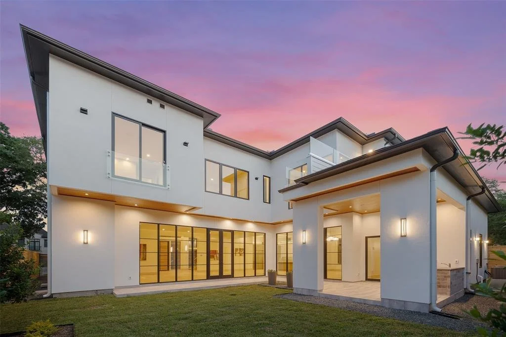 Modern white residential house with large glass windows and doors, exterior lighting, and a well-maintained lawn, under a colorful sunset sky.