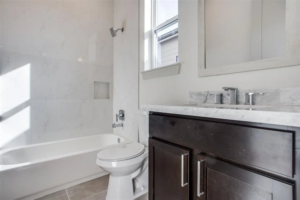 Bathroom with a bathtub, toilet, dark wood vanity with a marble countertop, and a mirror above the sink. There is a window next to the mirror, and the walls and bathtub surround are covered in white marble tiles.