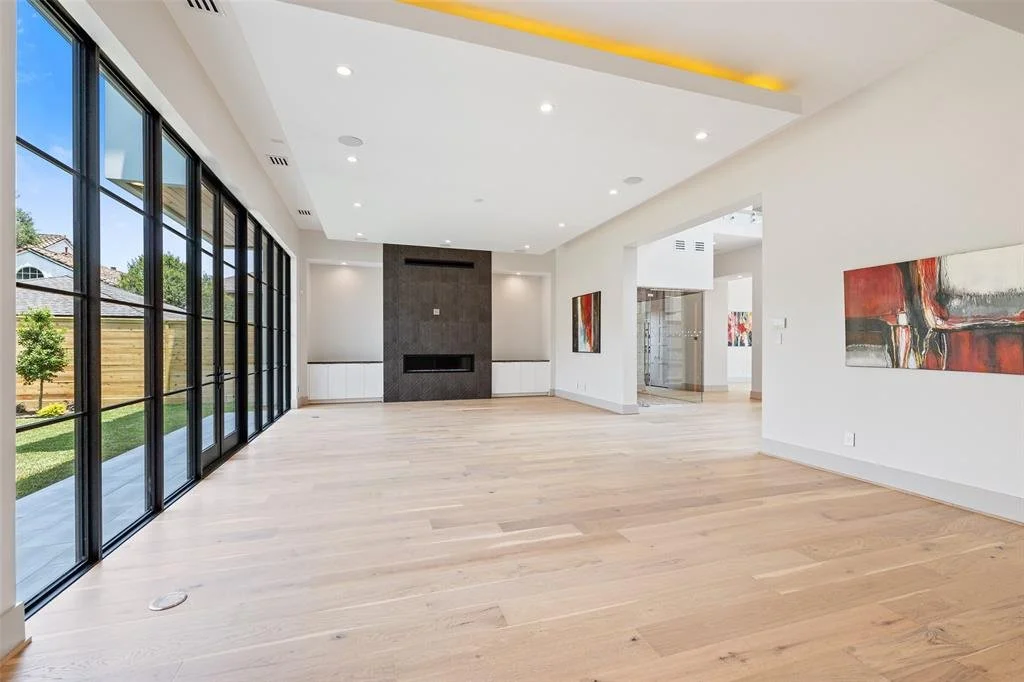 Spacious living room with large windows, light wood flooring, a modern fireplace with a dark surround, and abstract paintings on white walls.