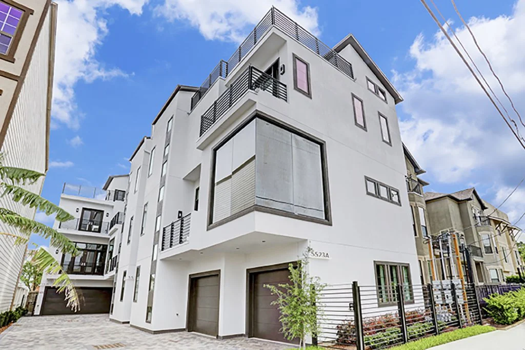 Modern white multi-story residential building with balconies, large windows, and garage doors, set against a blue sky with clouds.