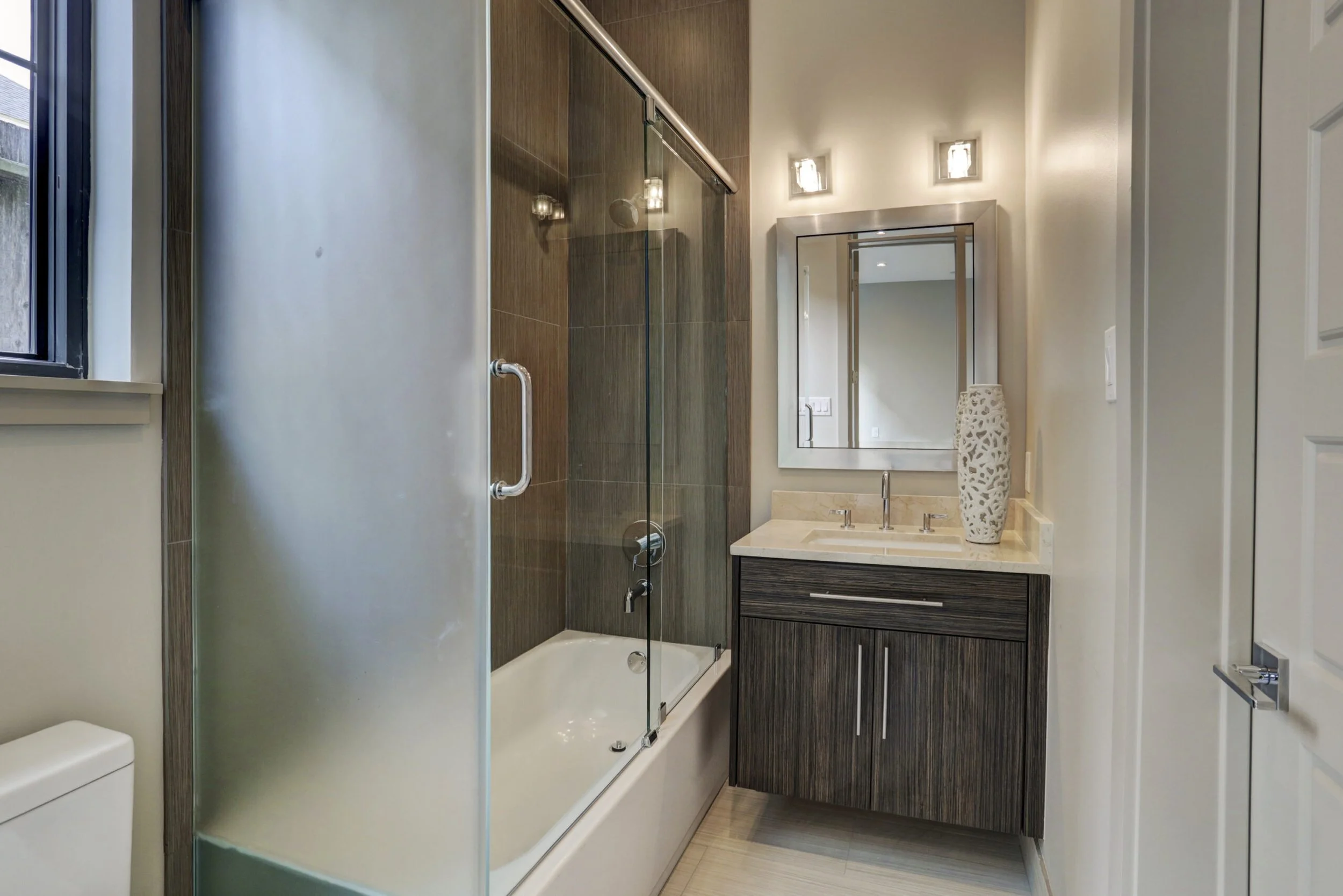 Modern bathroom with bathtub and glass shower door, dark wood vanity with a white countertop and a decorative vase, mirror, bathroom lighting, window with natural light, and white walls.