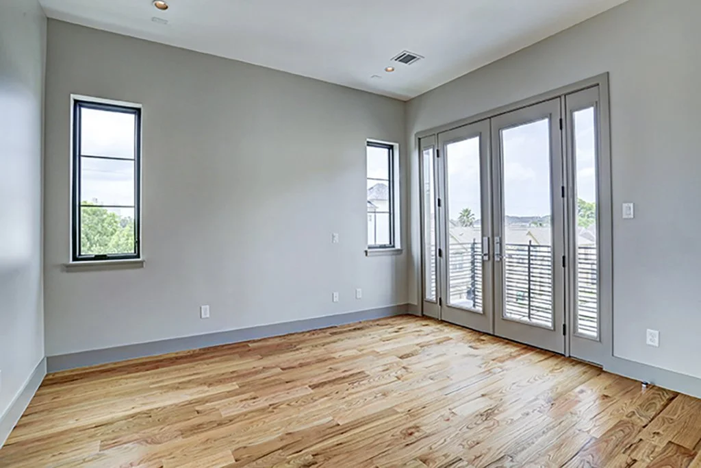 Empty room with hardwood floors, light gray walls, two small windows, and glass doors leading to a balcony.