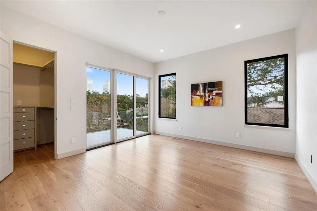 Empty living room with hardwood floors, white walls, large sliding glass doors leading to a balcony, and two windows with black frames showing trees and neighborhood views; artwork hanging on the wall; door leading to a closet.