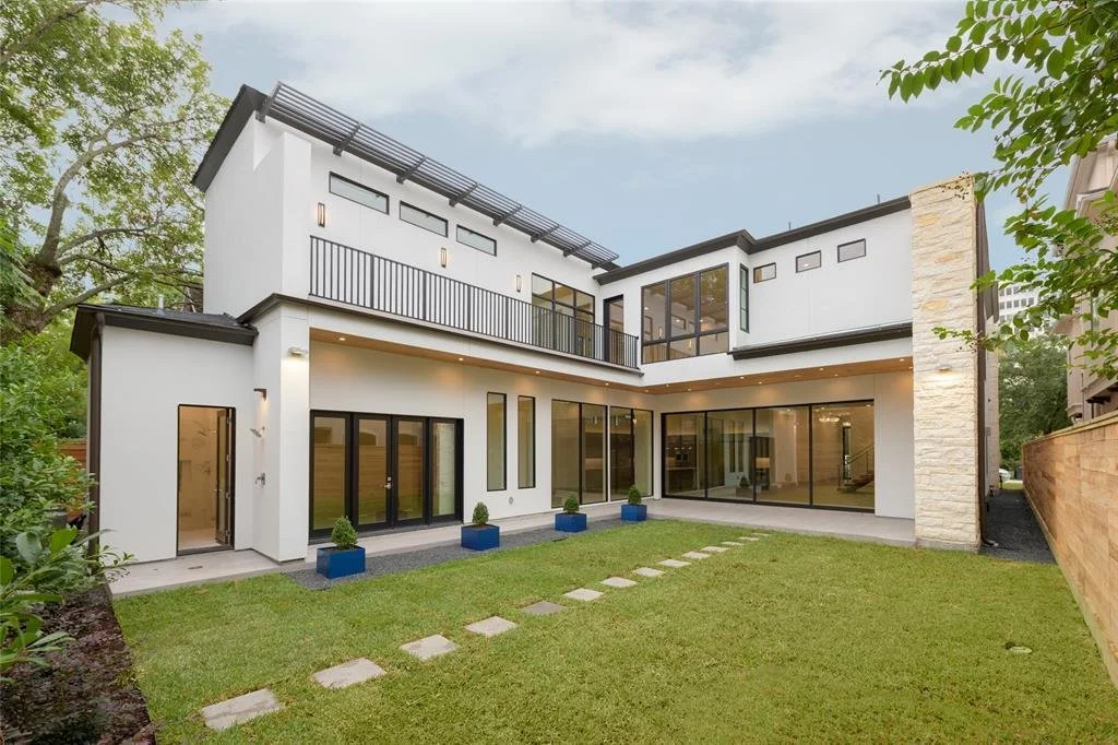 Modern two-story house with large glass windows and sliding doors, surrounded by a green lawn with stepping stones, some potted plants, and trees nearby.