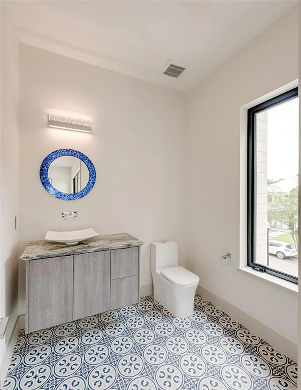 Modern bathroom with white walls, a large window, a wooden vanity with a marble countertop, a contemporary white toilet, a round blue mosaic mirror, and patterned floor tiles.