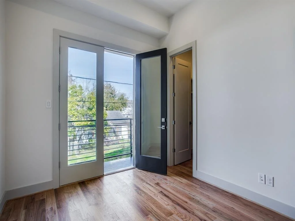 Empty room with hardwood floors, white walls, glass door leading to balcony, and an open black door to a closet or another room.
