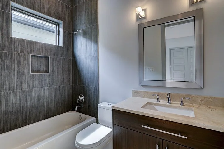 Modern bathroom with bathtub, toilet, dark wood vanity with a marble countertop, large mirror, and a small window