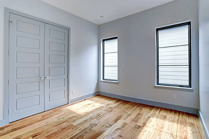 Empty room with light blue walls, wooden floor, two windows with black frames, and a closed double door.