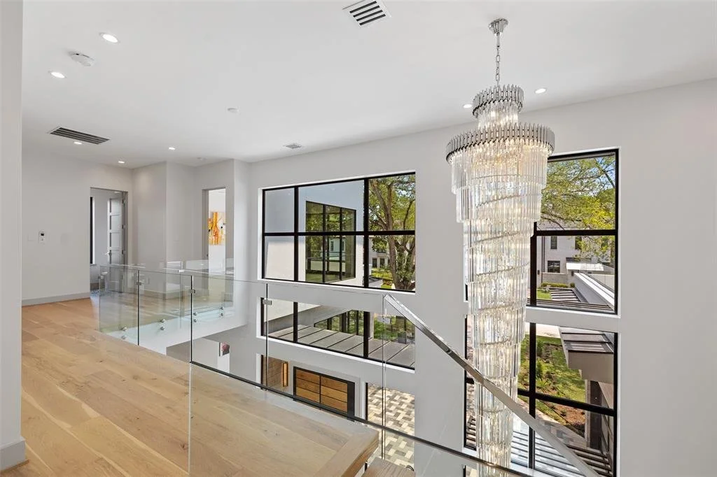Interior view of a modern, white-walled home with a large crystal chandelier, hardwood floors, and expansive windows showing trees and neighboring buildings.