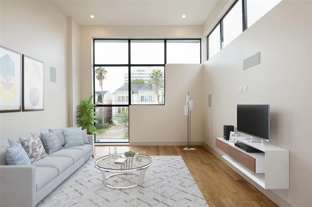 Bright living room with large windows, white walls, a white couch with patterned pillows, a glass coffee table, a white media console with a TV, a tall floor lamp, a green plant, and framed art on the walls.