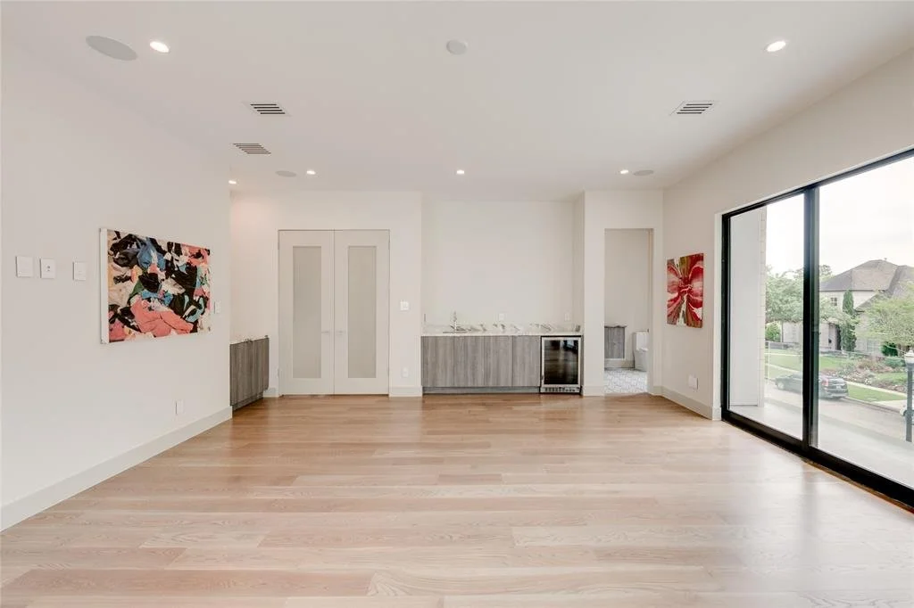 Empty living room with light-colored hardwood floors, white walls, two colorful paintings, large sliding glass doors leading outside, and a small kitchenette in the background.
