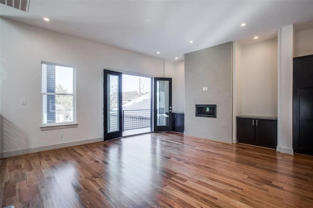 Empty living room with hardwood floors, white walls, and a gray accent wall with a modern fireplace. There are two windows and a glass door leading to a balcony, with black frames and trim, allowing natural light into the room.