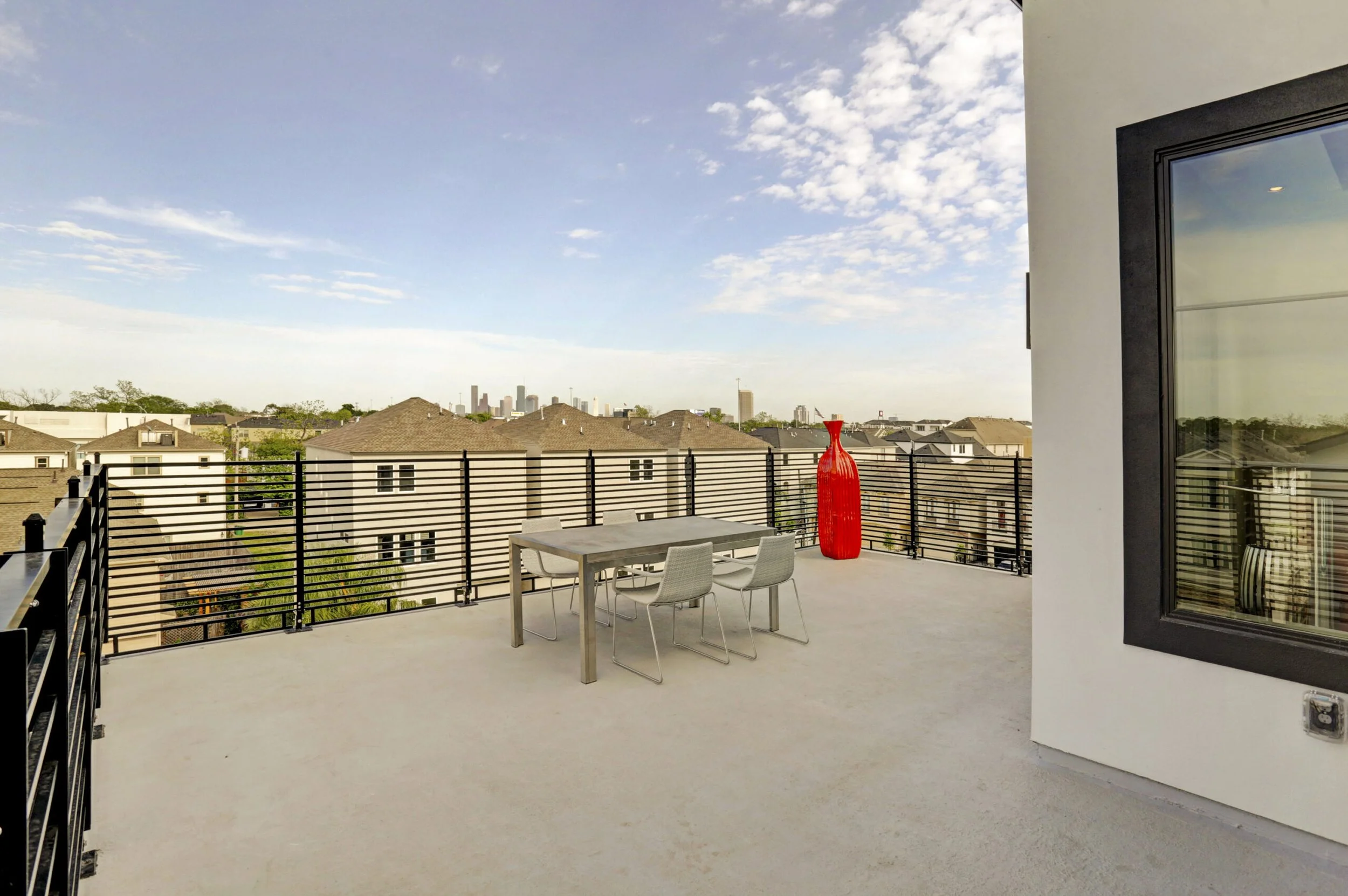 Empty balcony with a table, four chairs, a large red vase, and a black railing overlooking a residential neighborhood with a city skyline in the distance under a partly cloudy sky.