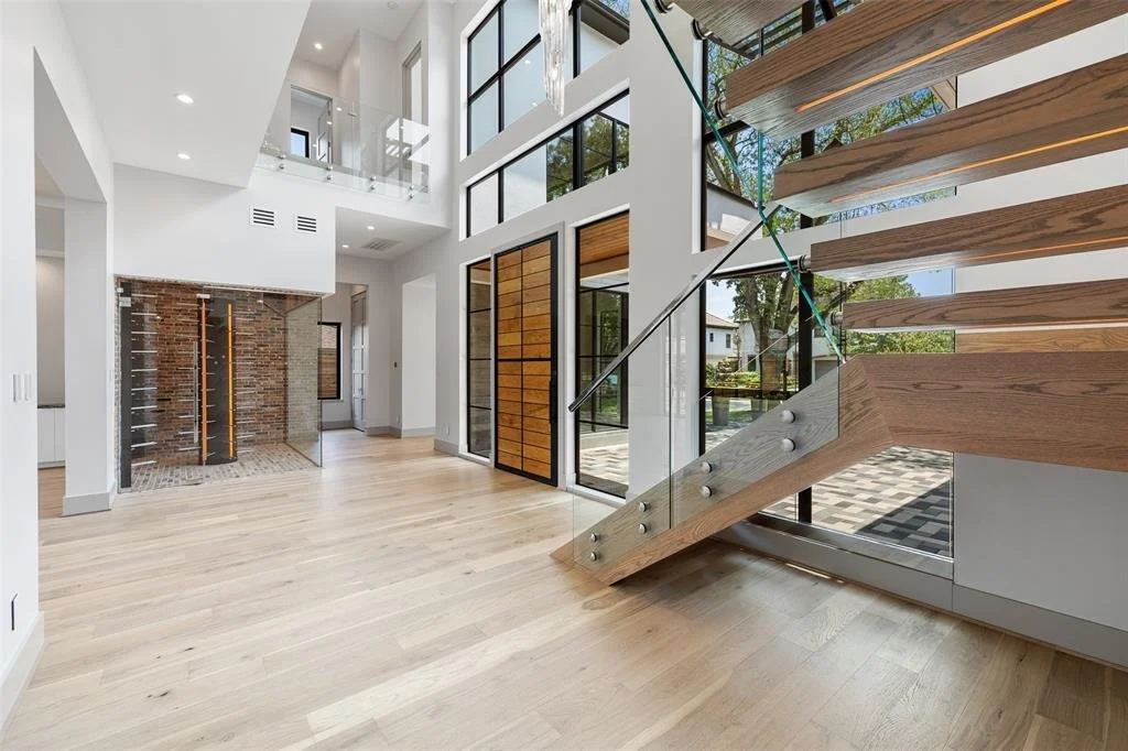 Interior of a modern, spacious house featuring a wooden staircase with glass railing, large windows, and a brick fireplace.