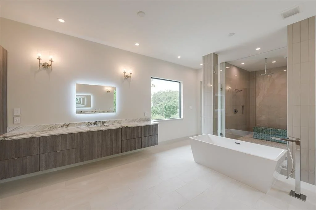 Modern bathroom with a freestanding bathtub, a large shower area, a long vanity with a marble countertop, a rectangular mirror with backlighting, two wall sconces, and a window showing greenery outside.
