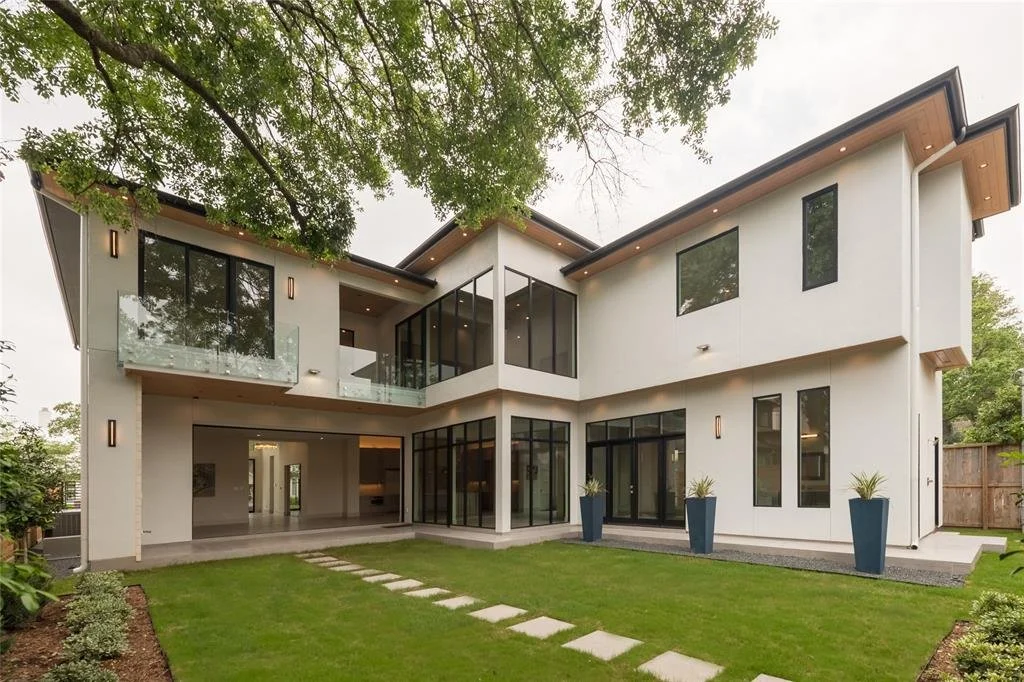 Modern two-story house with large glass windows and sliding doors, surrounded by a neatly manicured lawn and potted plants, with a wooden fence and trees in the background.