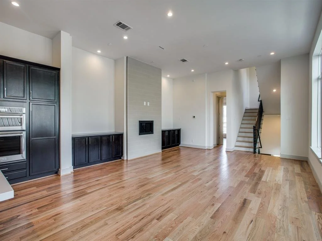 Empty modern living room with hardwood floors, black kitchen cabinets, a fireplace, staircase, and large window.
