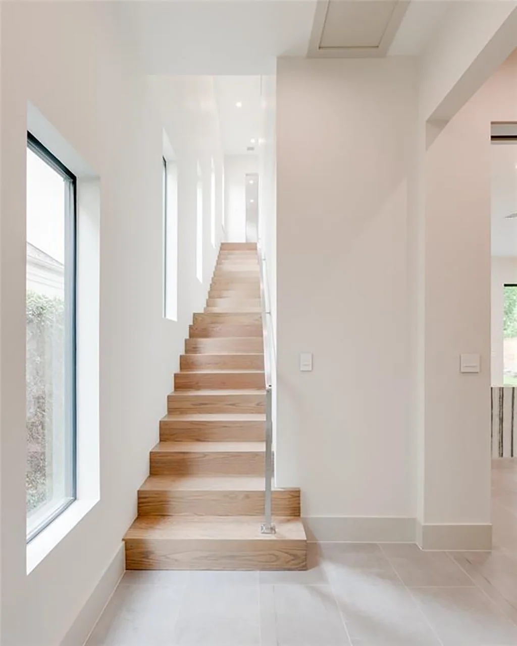 Modern interior staircase with wooden steps, white walls, and large windows allowing natural light.