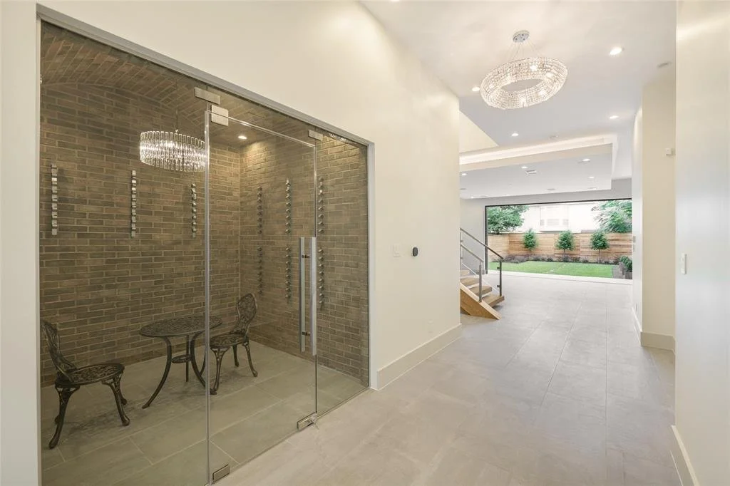 Modern interior space with a glass-walled room featuring brick walls, a small round table, and two chairs; an open area with a chandelier, recessed lighting, and large windowed door leading to a backyard with trees and plants.