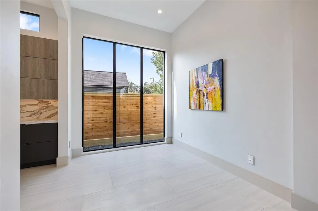 Empty room with white walls, light-colored flooring, a large sliding glass door opening to a small backyard with a wooden fence, and a colorful abstract painting on the wall.