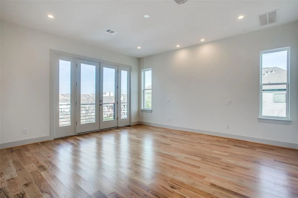Empty room with hardwood floors, white walls, three large glass doors leading to a balcony, and two windows, with ceiling lights.