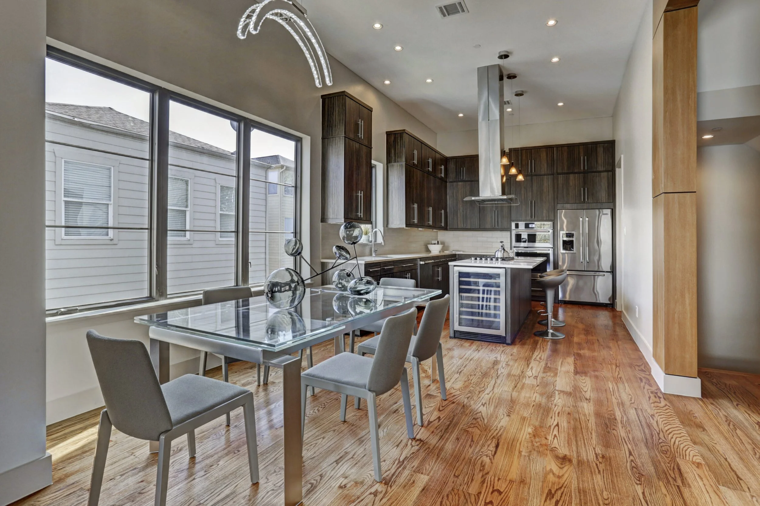 Modern kitchen and dining area with large windows, wooden flooring, dark cabinets, stainless steel appliances, a glass dining table with chairs, and decorative glass vases on the table.