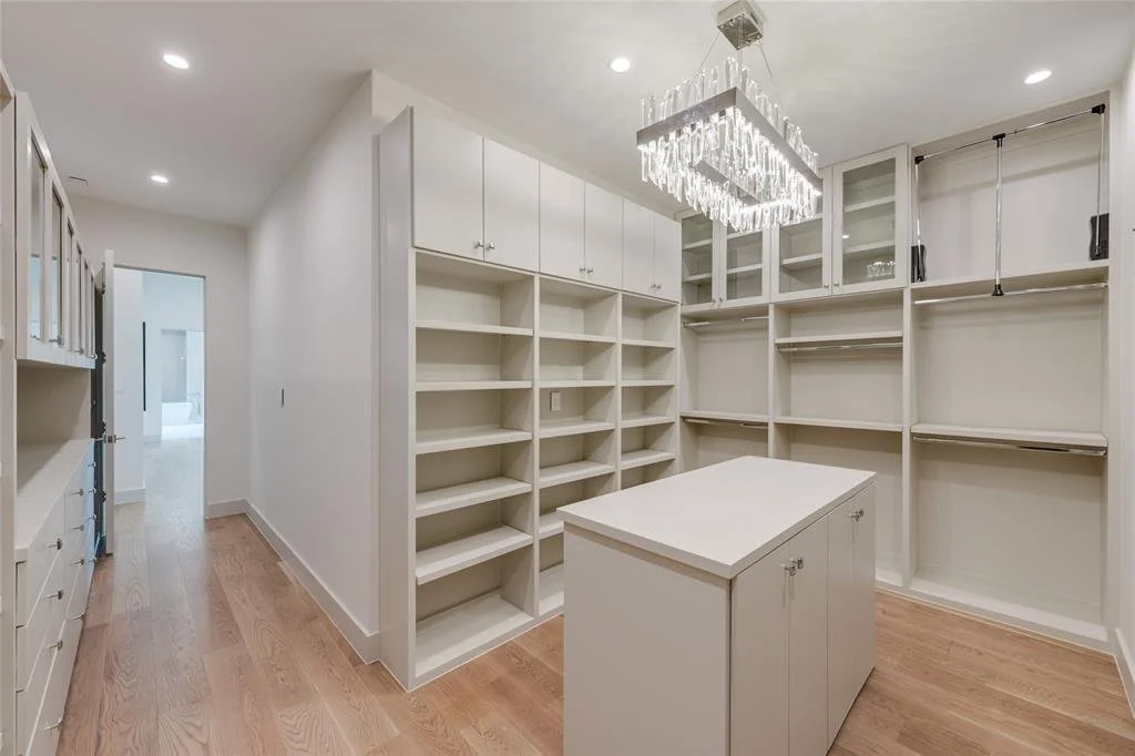 A walk-in closet with white cabinetry, open shelves, and a center island, illuminated by a large rectangular chandelier with hanging crystals.
