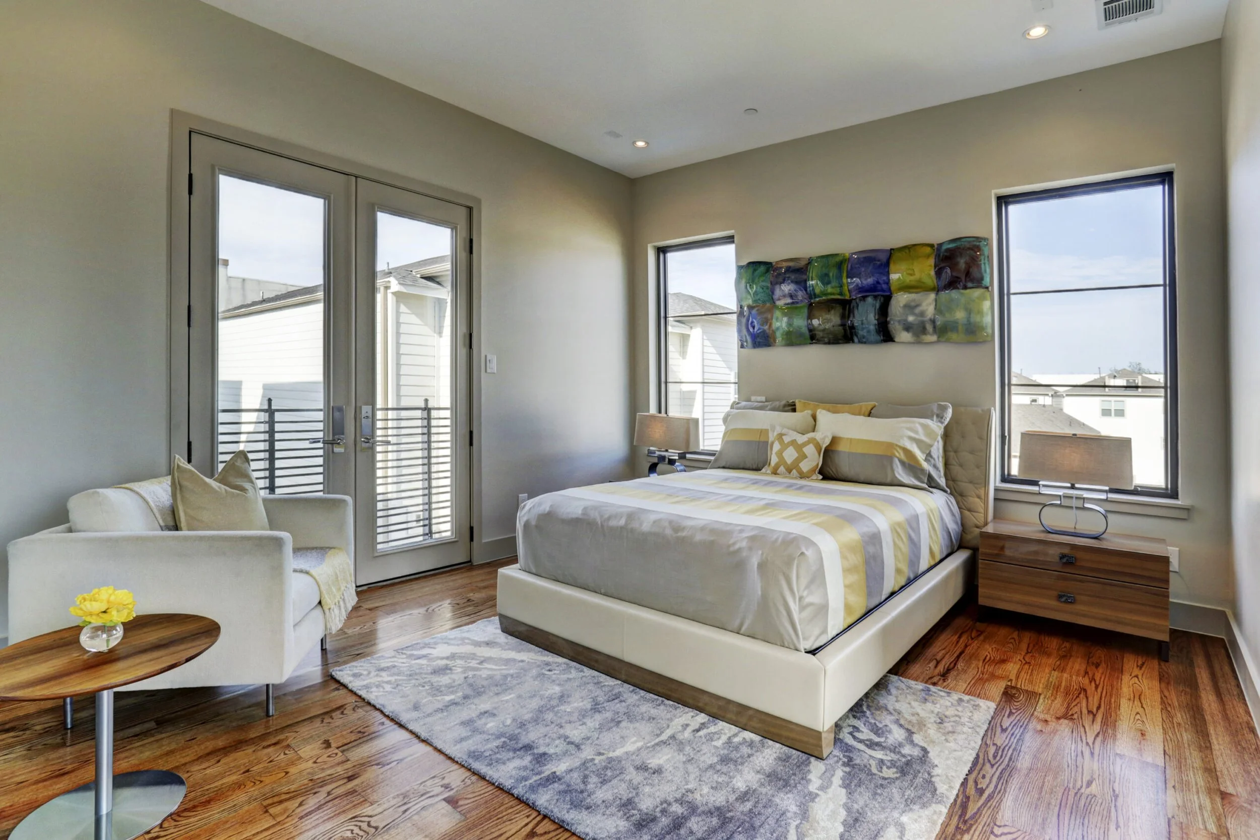 A modern bedroom featuring a bed with yellow and gray striped bedding, a wooden nightstand, lamps, a white armchair with a yellow pillow, and a wall hanging abstract art piece, with hardwood floors and a window and glass door leading to a balcony.