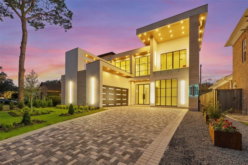 Modern two-story house with large glass windows and a driveway at dusk, illuminated with exterior lighting.