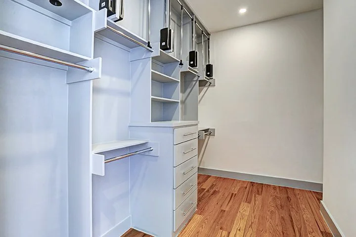 Empty walk-in closet with white shelving and drawers, wooden floor, and recessed ceiling lights.