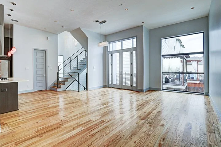 Empty living room with large windows, hardwood floors, and a staircase leading upstairs.