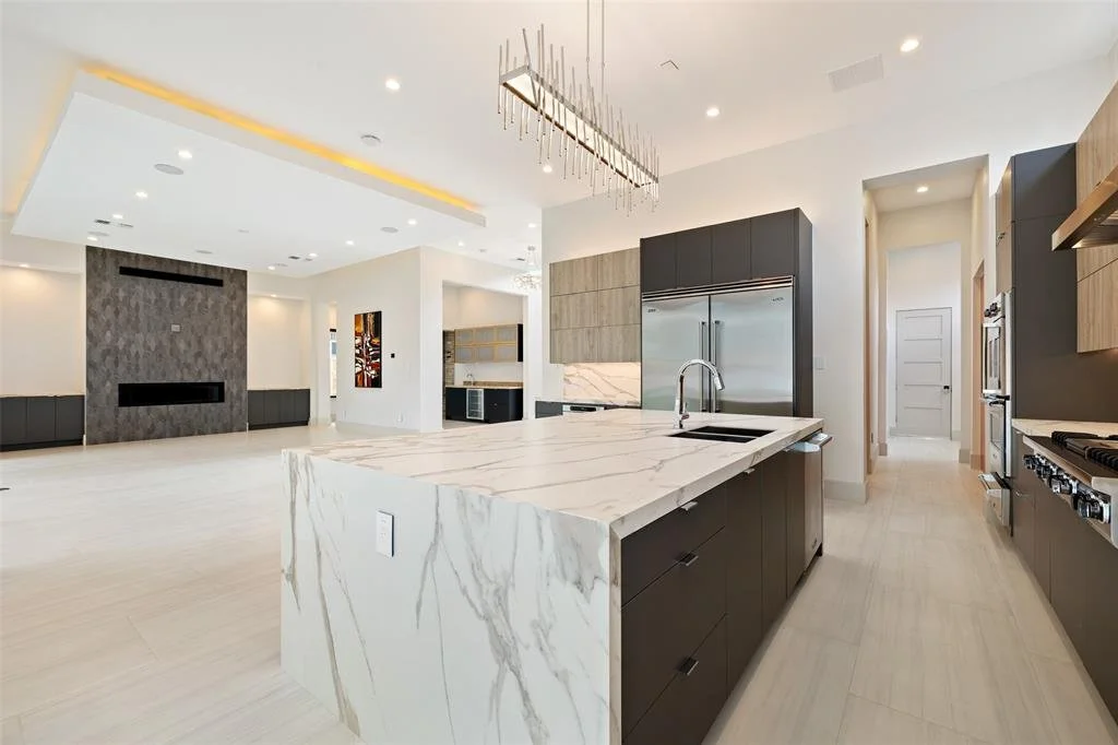 A modern, open-concept kitchen and living space with white marble island, stainless steel refrigerator, black and wood cabinets, and minimalistic decor.