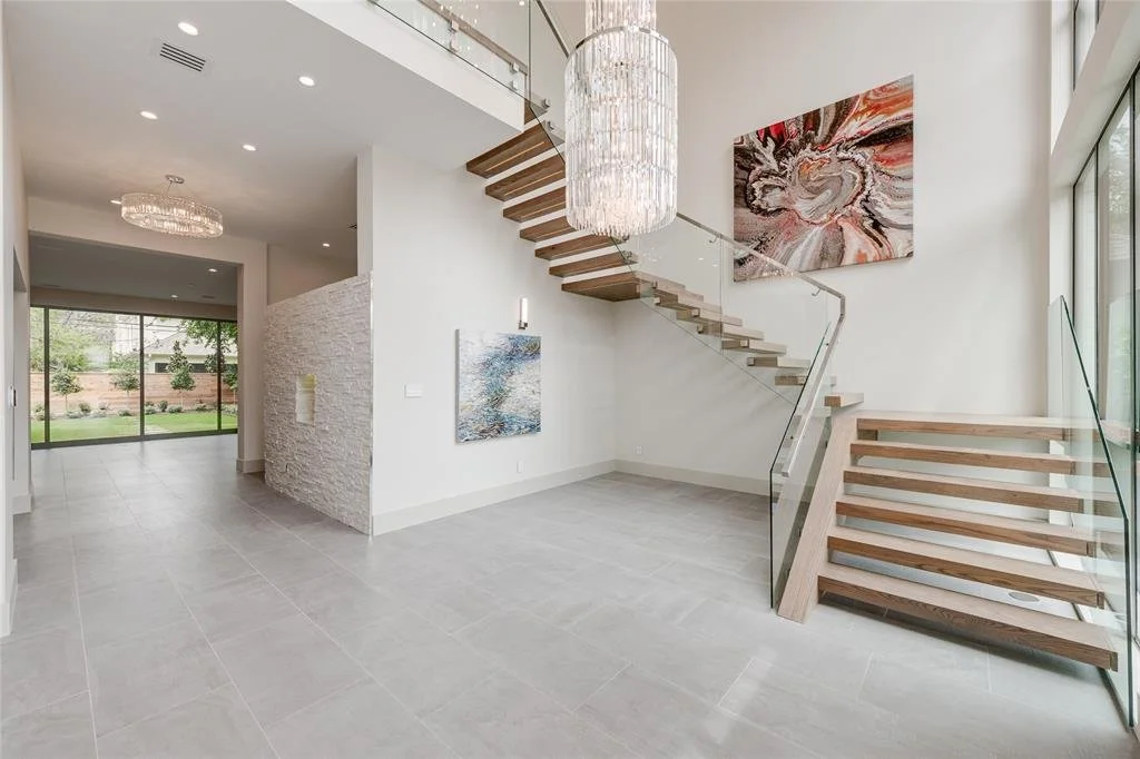 Modern open-concept living space with a staircase, large window, artwork, and a chandelier, with a view of a backyard.