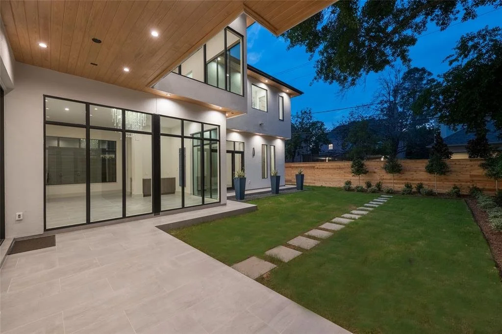 Modern house backyard with sliding glass doors, a wooden ceiling over the patio, a well-maintained lawn with a stone pathway, and a wooden fence with small trees along it, during evening.
