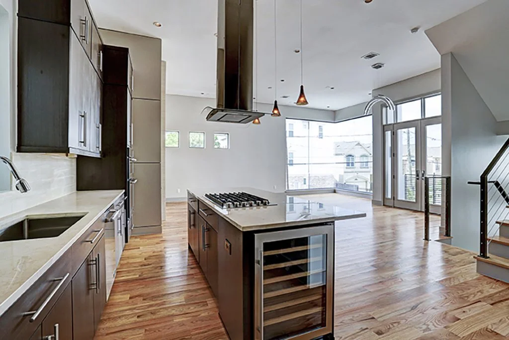 Modern open-concept kitchen and living area with wooden flooring, dark kitchen cabinets, an island with a built-in wine cooler, stainless steel appliances, large windows, and a staircase with metal railings.