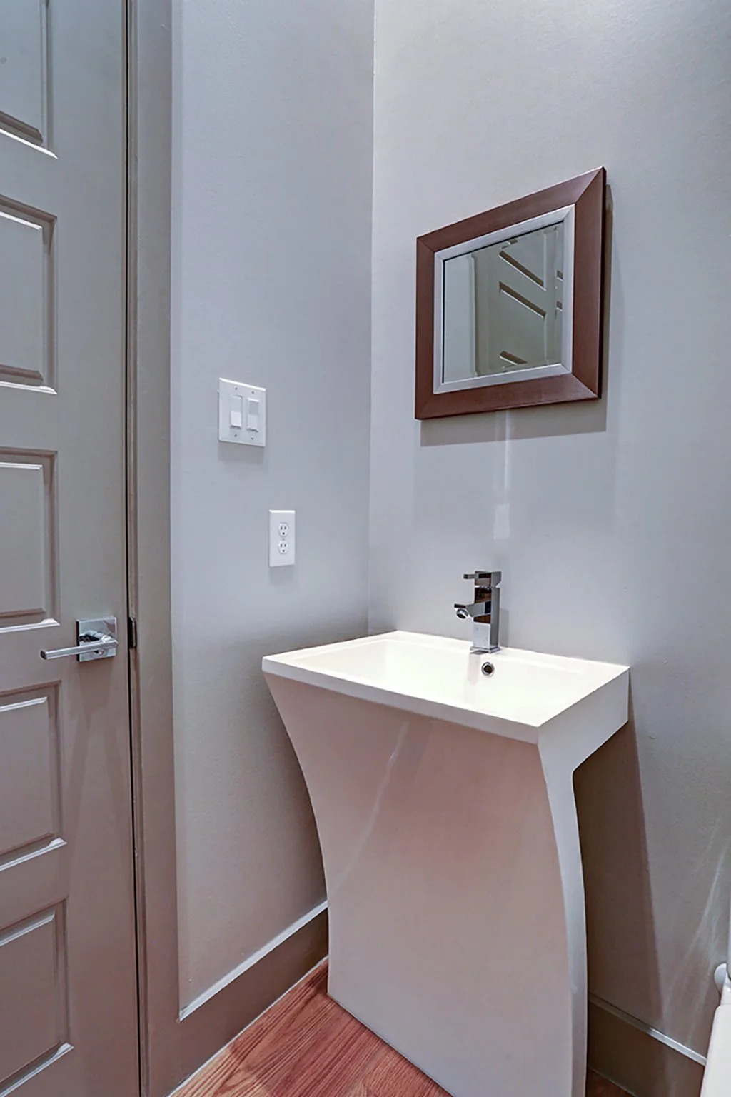 A small white triangular sink with a chrome faucet is mounted on the wall in a corner of a bathroom or utility space. Above the sink, a rectangular mirror with a wooden frame is hung on the wall. To the left, there is a light switch and an electrical