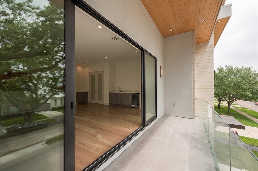 Modern balcony with glass railing overlooking green trees and a residential street, with a sliding glass door leading inside to an empty room with wood flooring.