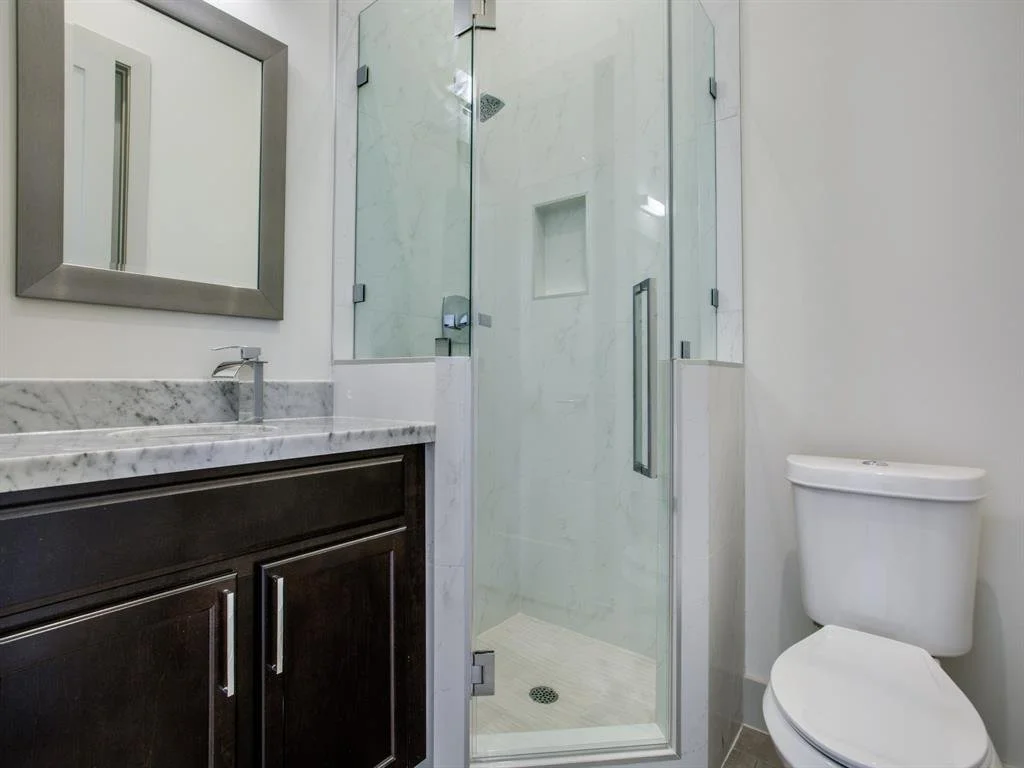 Modern bathroom with a glass shower enclosure, a dark wooden vanity with a marble countertop, a rectangular mirror with a silver frame, and a white toilet.