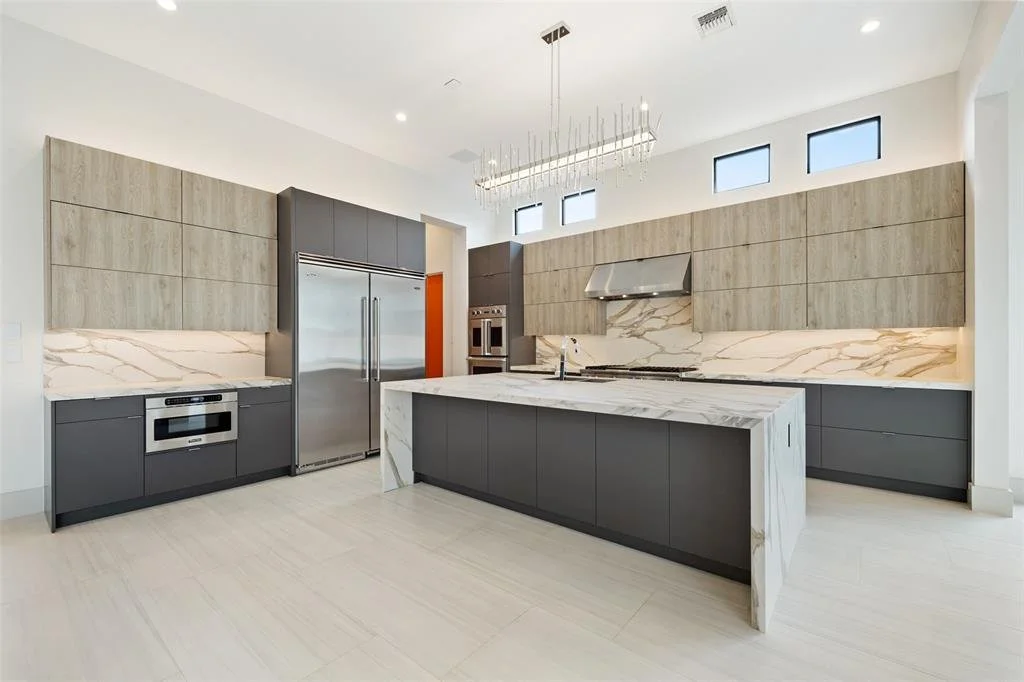Modern kitchen with gray lower cabinets, light wood upper cabinets, marble backsplash, kitchen island, stainless steel appliances, and high windows.