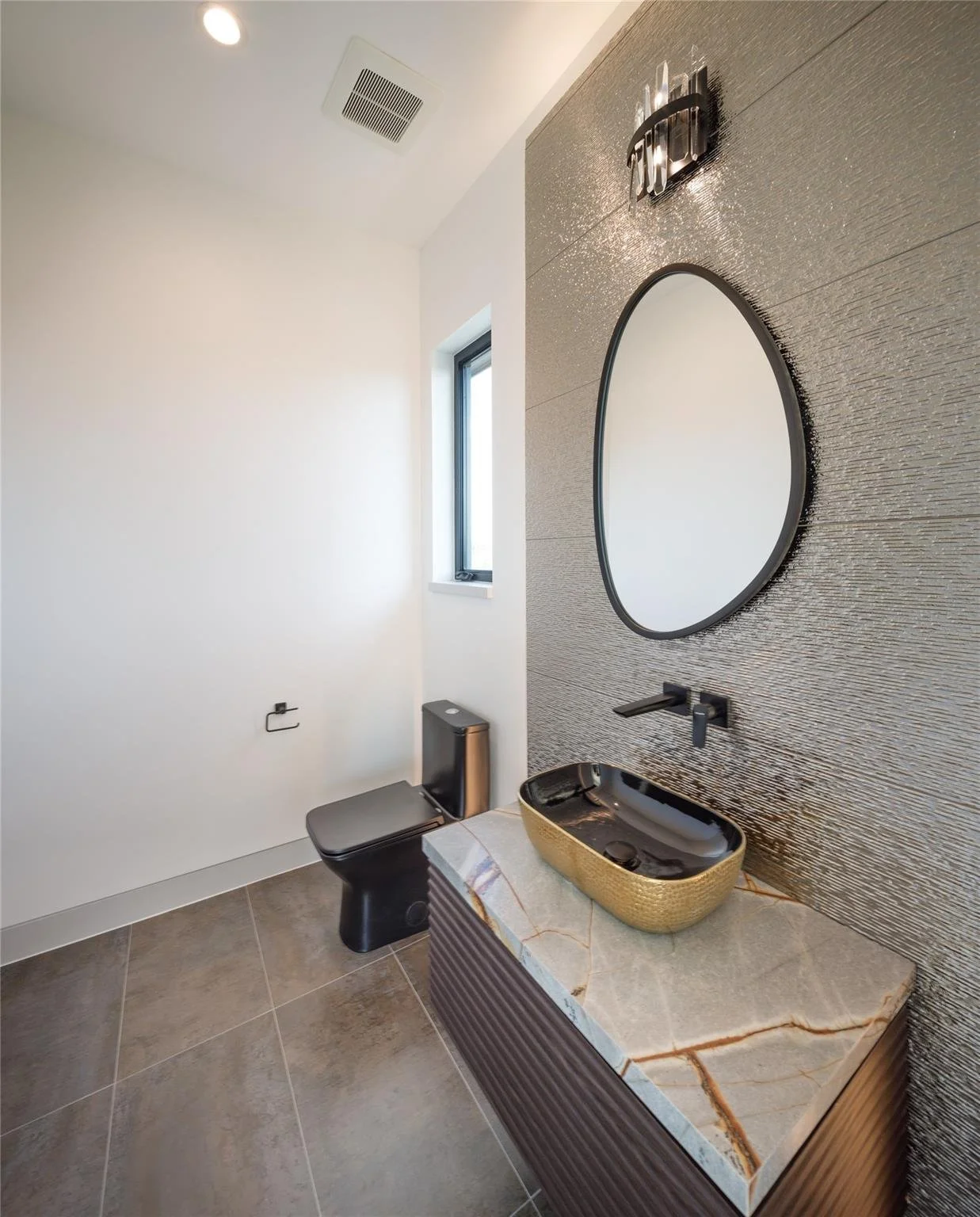 Modern bathroom with a marble countertop, black vessel sink, round mirror, black wall-mounted faucet, wall sconce, black toilet, and a small window.