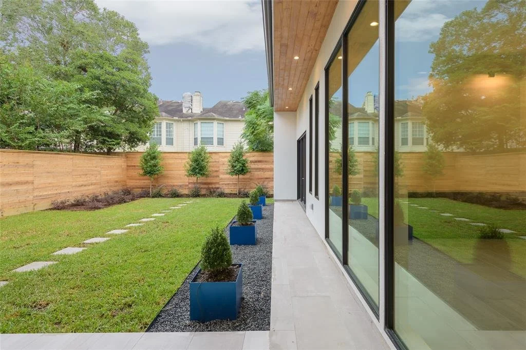 A backyard lawn with a wooden privacy fence, small trees in blue planters near a modern house with large glass windows and a wooden ceiling overhang.