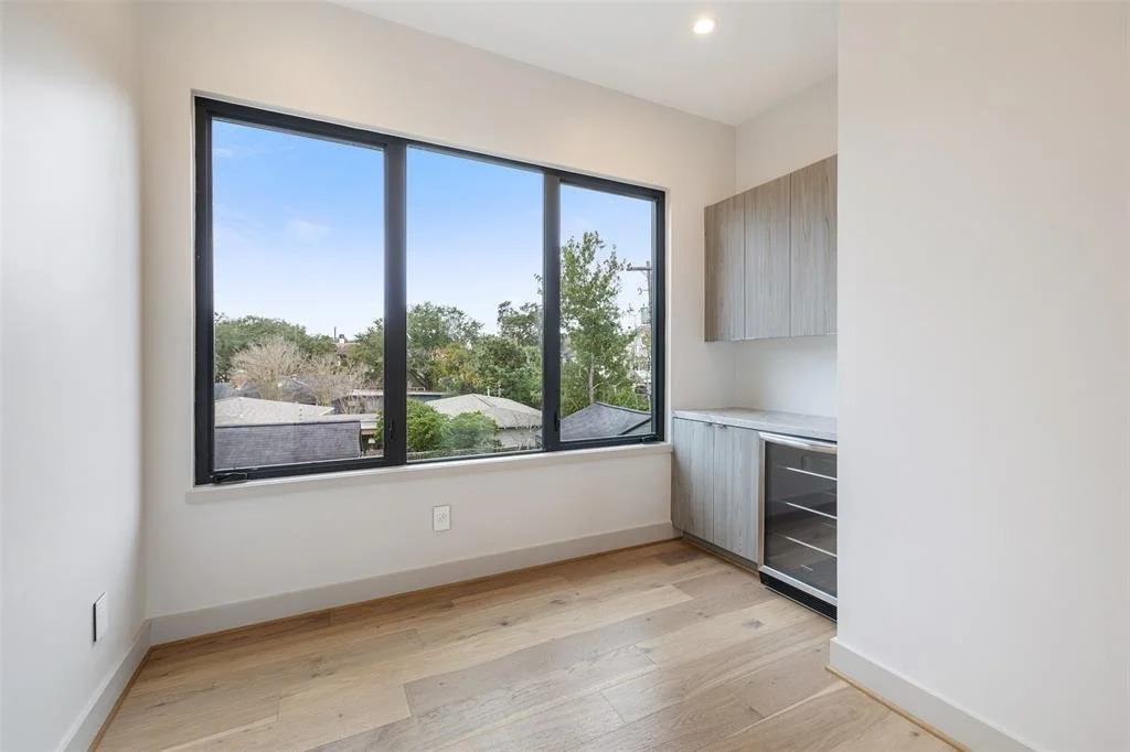 Empty room with large window showing a view of trees and houses, light-colored wooden cabinets, and a small wine cooler.