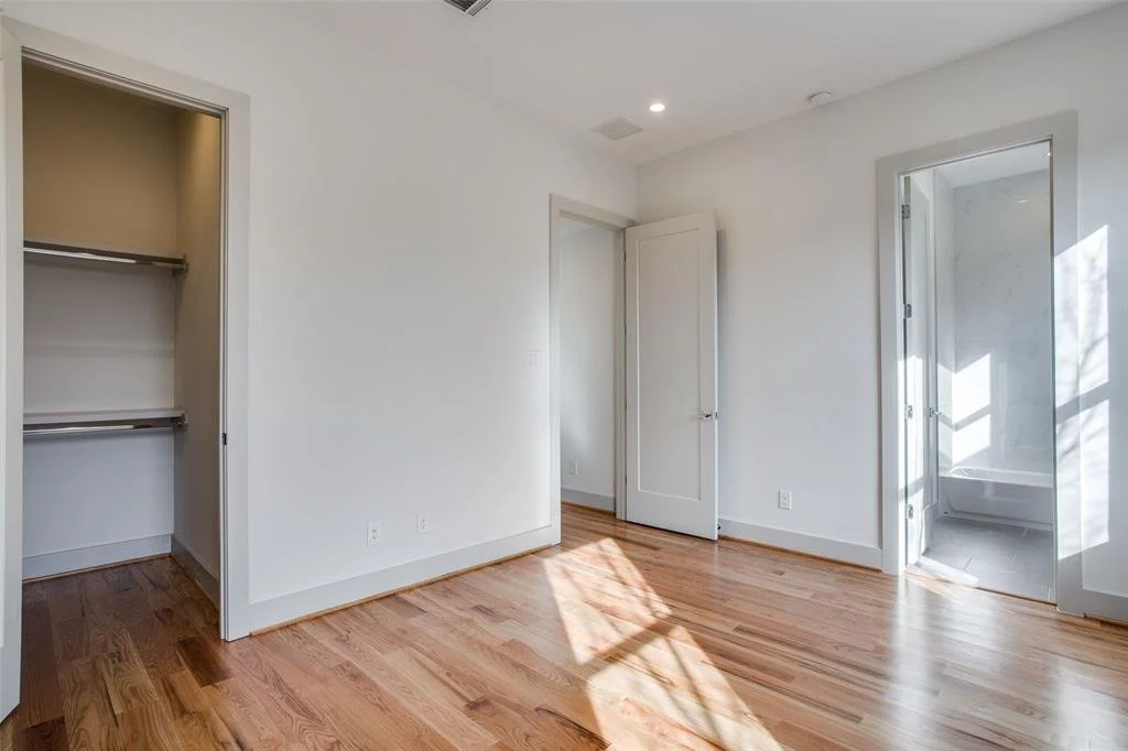 Empty bedroom with hardwood floors, open closet, and a door leading to a bathroom with a bathtub. Sunlight casting shadows on the floor.