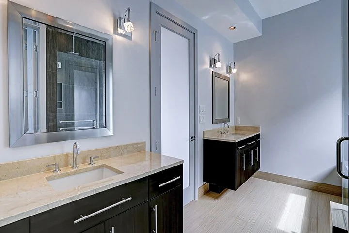 Bathroom with two black vanities with beige countertops, mirrors, and wall-mounted lights, next to a glass shower door.