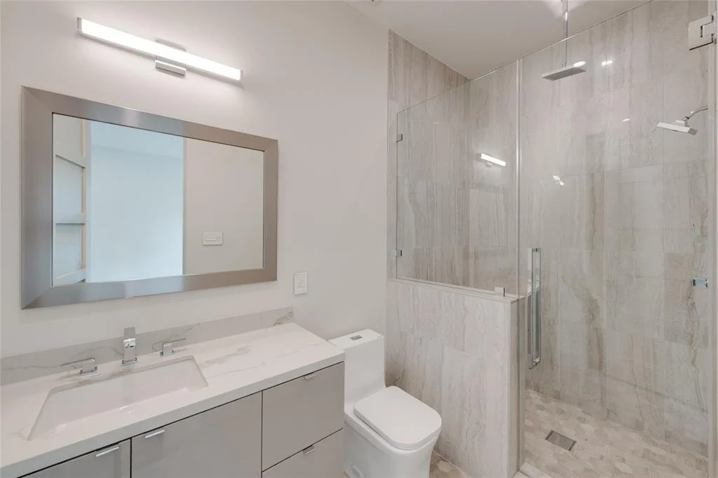 Modern bathroom with beige marble shower, glass door, white toilet, and gray vanity with large mirror.