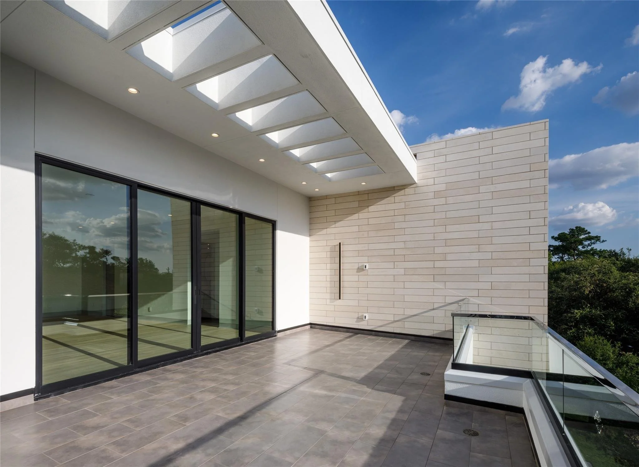 Modern balcony with sliding glass doors, tiled floor, white brick wall, and glass railing overlooking trees and blue sky.