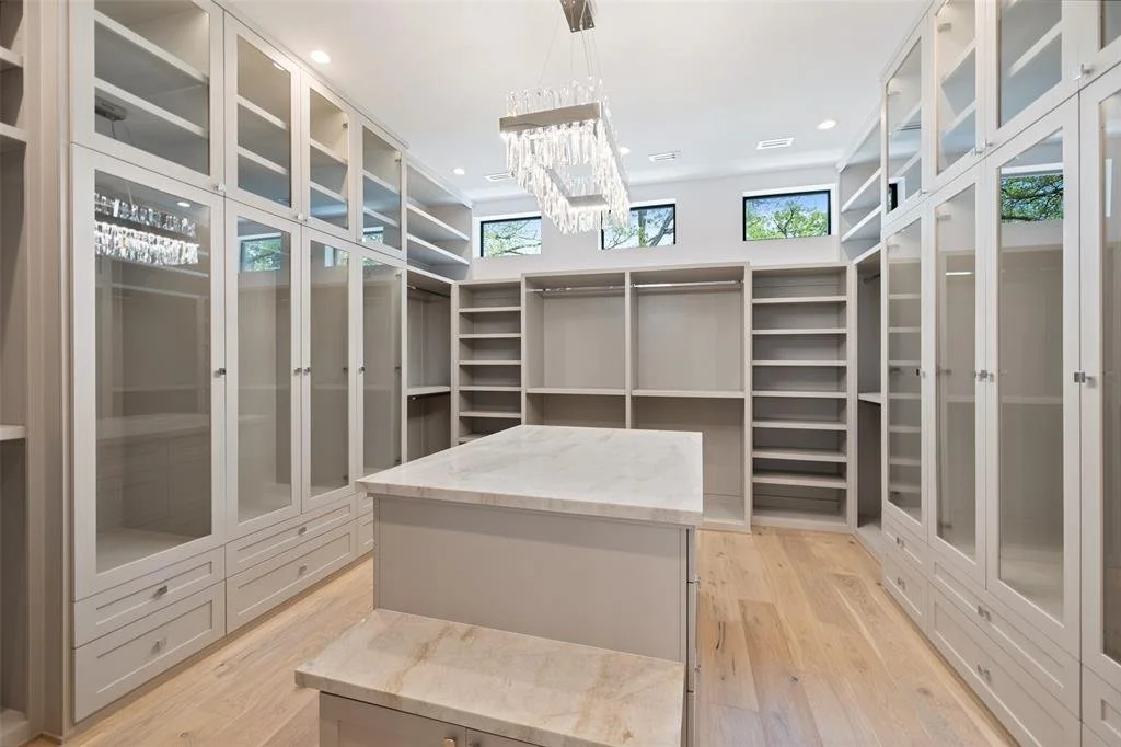 Empty walk-in closet with white cabinets and shelves, a central island, wooden flooring, and windows near the ceiling.
